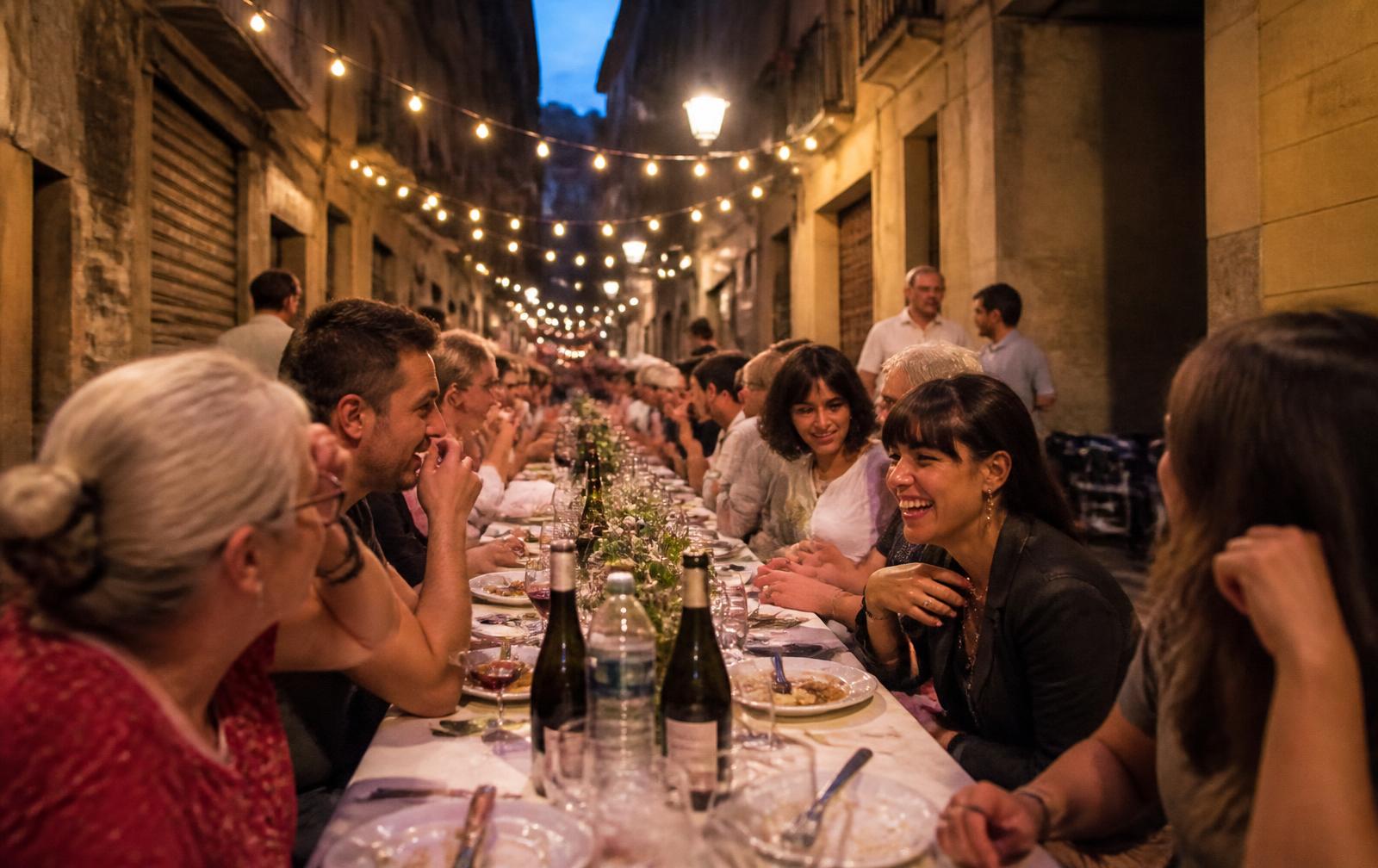 A long shared dinner table on a city street at dusk, lit by string lights